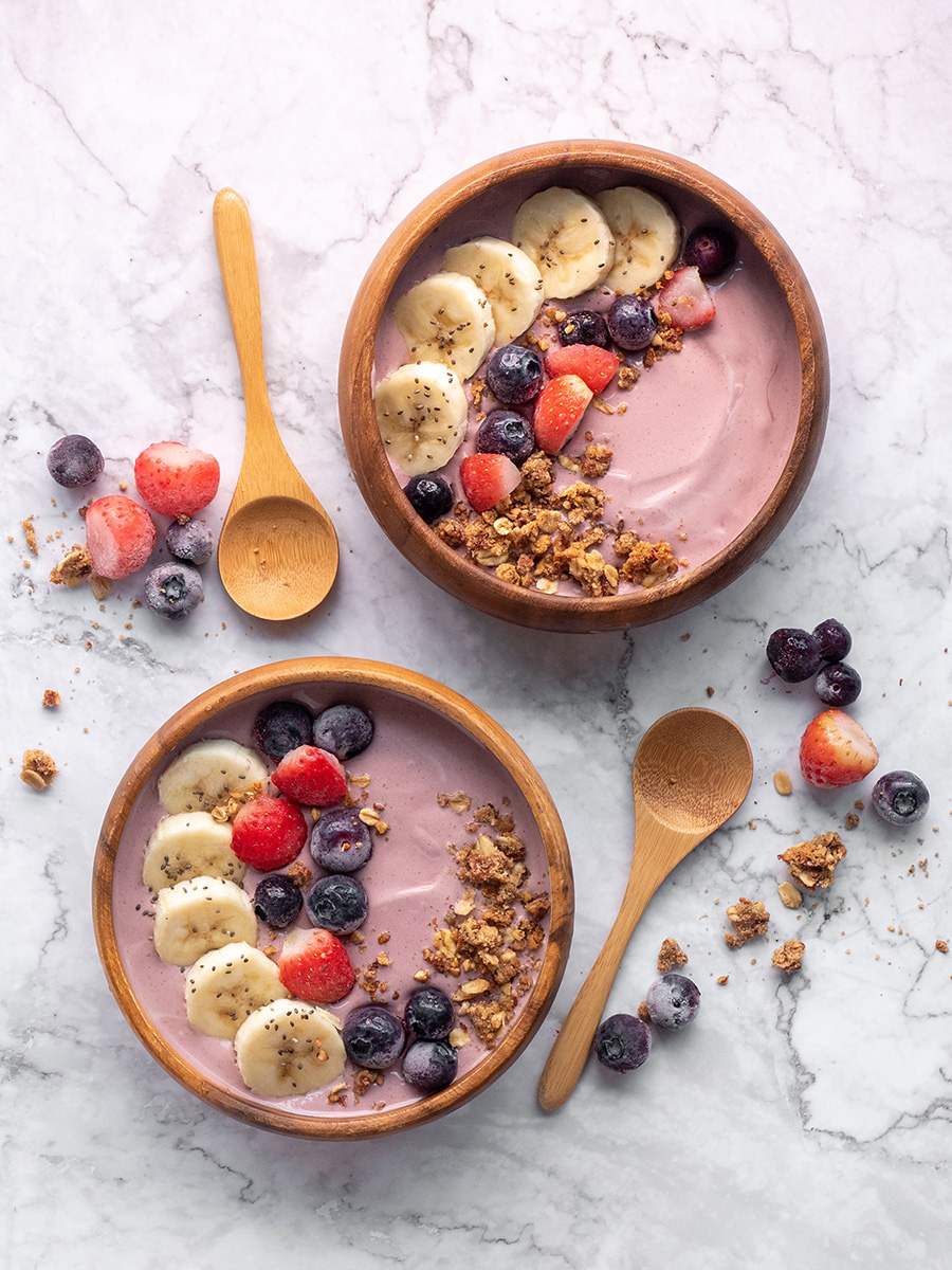 Two bowls of yoghurt topped with bananas, granola and mixed berries placed beside wooden spoons on a marble table