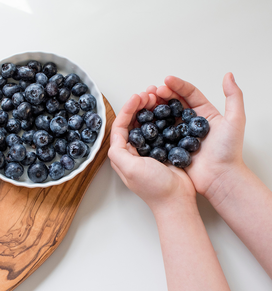 A child taking a handful of berries from a bowl filled with blueberries